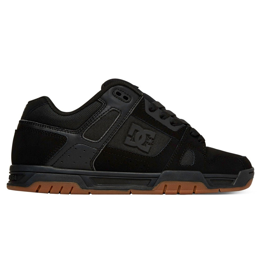 The DC Shoes Men’s Stag Shoes are shown in side view against a white background, featuring a black low-profile design with mesh and suede uppers, black laces, perforated panels, and a durable brown cupsole.