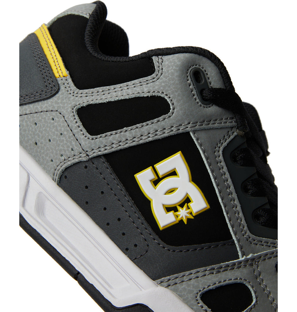 Close-up of the DC Shoes Men’s Stag sneaker in gray and black, featuring a prominent yellow and white DC Shoes logo on the side. Designed for extra comfort with an oversized puff tongue, this shoe also includes a durable cupsole and yellow accents near the heel.