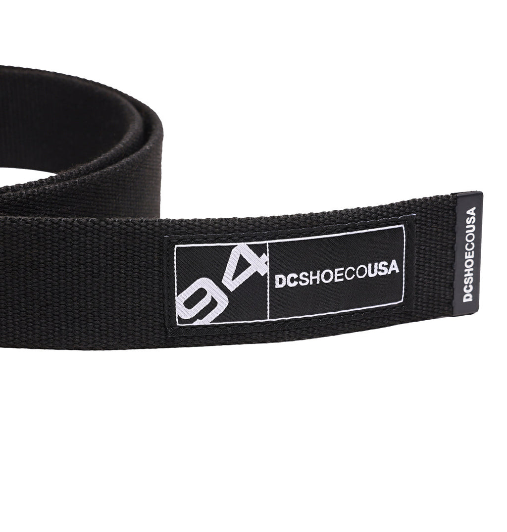 A close-up of the DC Stash Web Belt by DC Shoes, featuring a black adjustable webbed strap with a stitched label displaying “94 DCSHOECOUSA” in white text. The label is sewn onto the end of the belt, which is partially rolled up in the background.