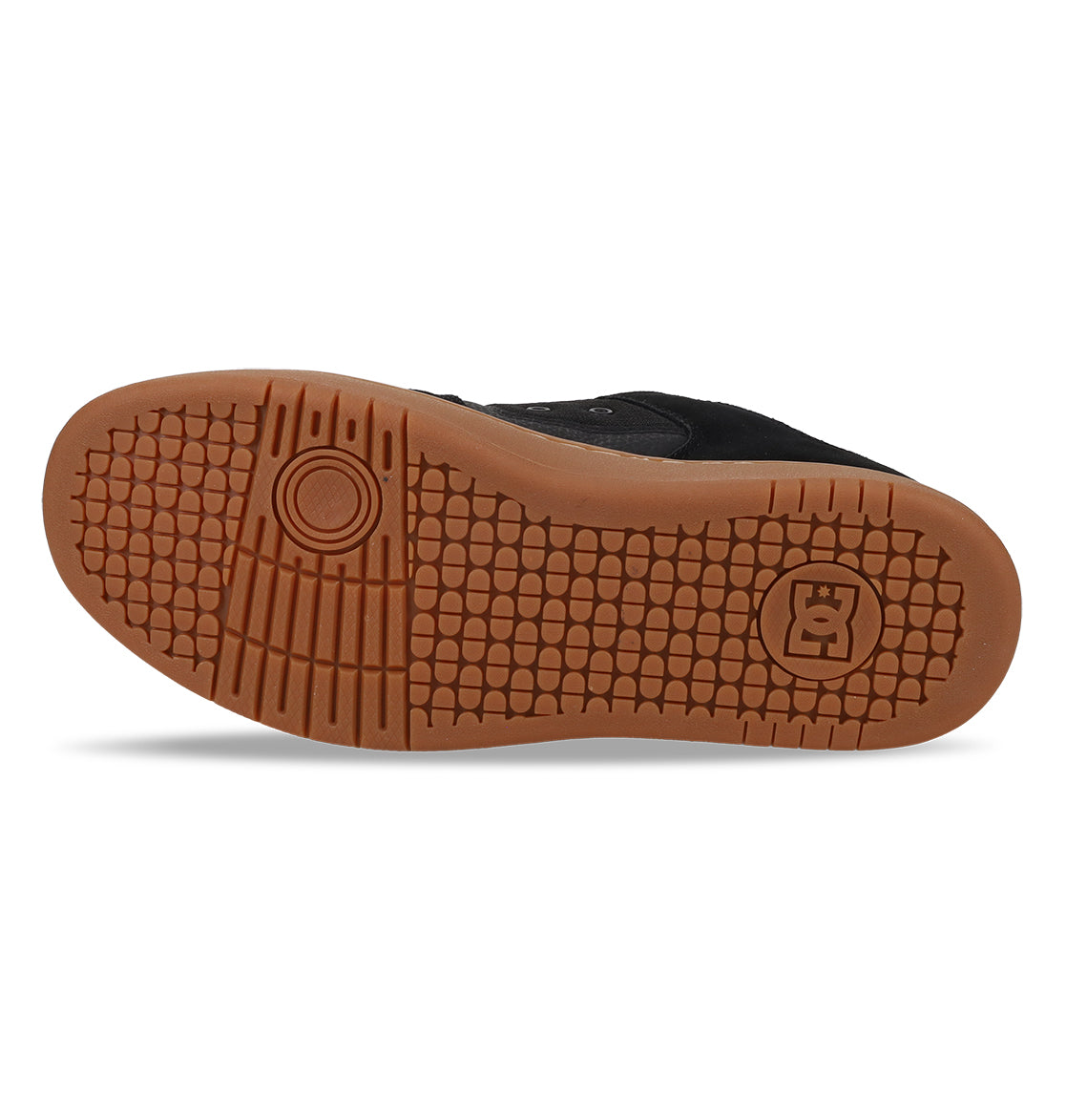 The image shows the brown rubber outsole of a black skate shoe, specifically the Men’s Manteca 4 Shoes from DC Shoes, featuring a textured grip pattern designed for modern comfort and two logos—one circular near the ball and one rectangular near the heel.