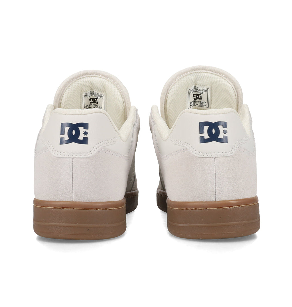 Two white Men’s Manteca 4 Shoes from DC Shoes, featuring tan soles, are displayed from the back. The performance-focused design highlights the iconic DC Shoes logo in dark blue on each heel, with insides and tags also partially visible.