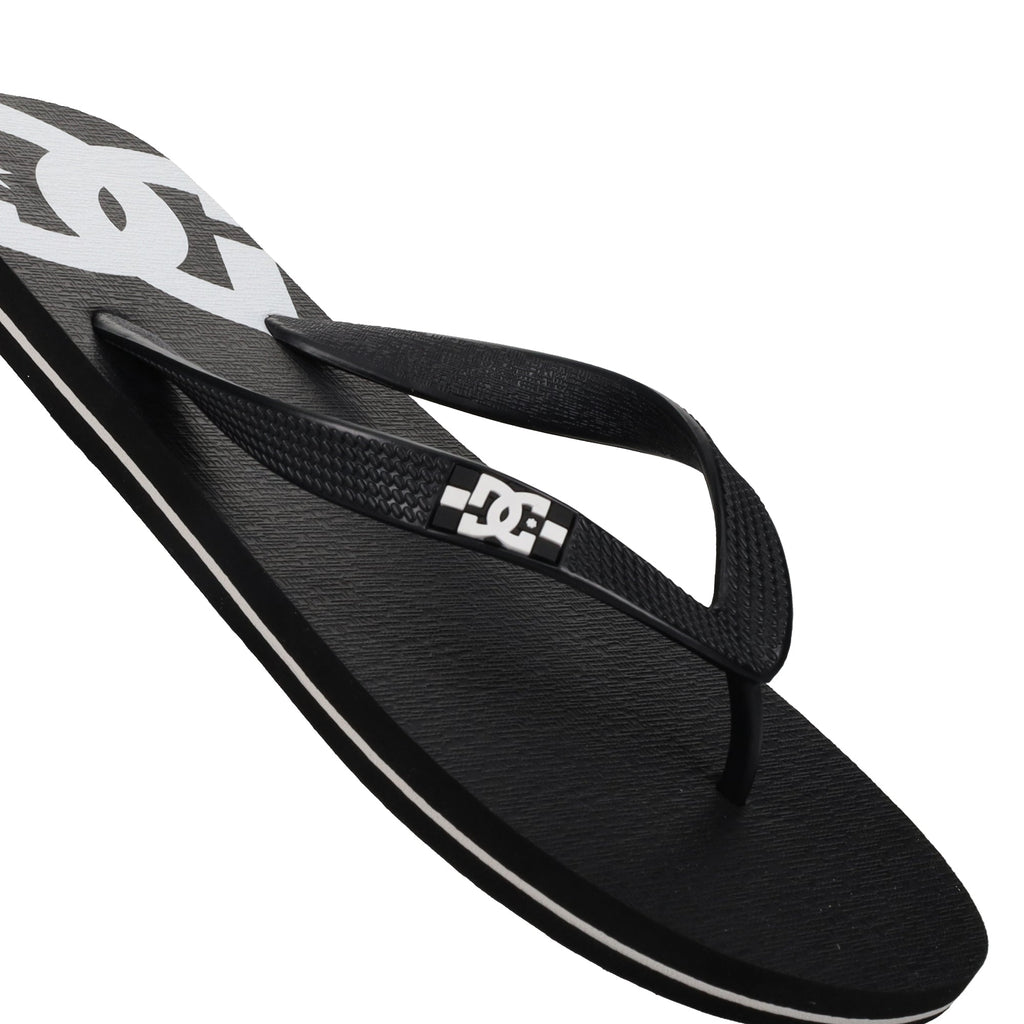 A close-up of the Men’s Spray Slides by DC Shoes showcases a black flip-flop sandal with a white DC logo on the strap and footbed, featuring a textured surface and white accents—these must-have slides are perfect for relaxing after a skate session.