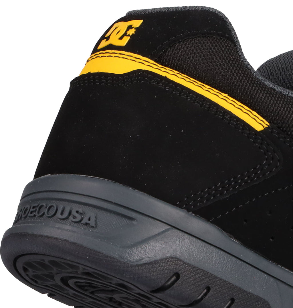 Close-up of the heel of the Men's Stag Shoes by DC Shoes. This black shoe features a yellow accent, DC logo on the back, and a durable dark gray cupsole embossed with SHOESCOUSA, capturing classic skateboarding style.