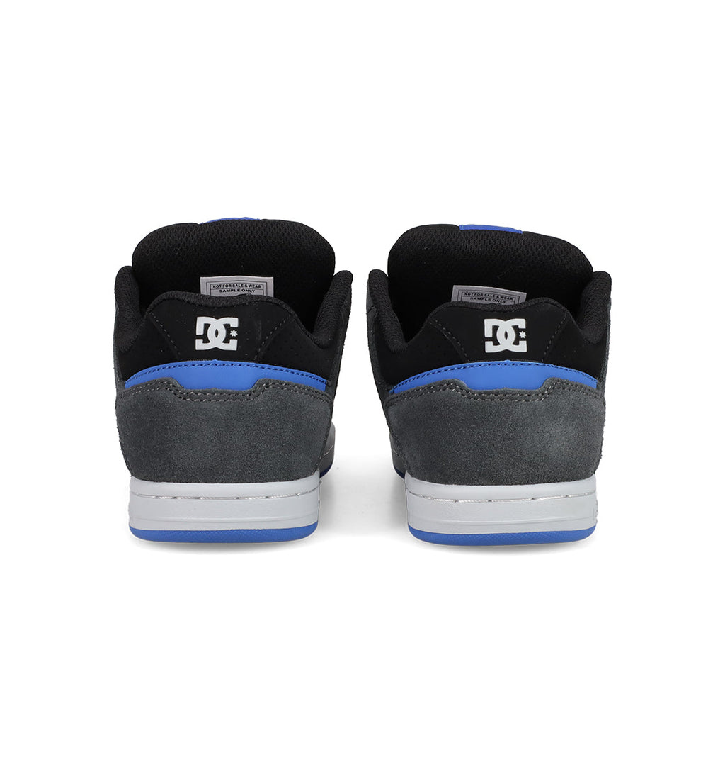 A pair of Kids' Stag Shoes by DC Shoes seen from the back, featuring black and grey suede uppers, blue trim, white soles, cupsole construction, and the DC logo on the heel.