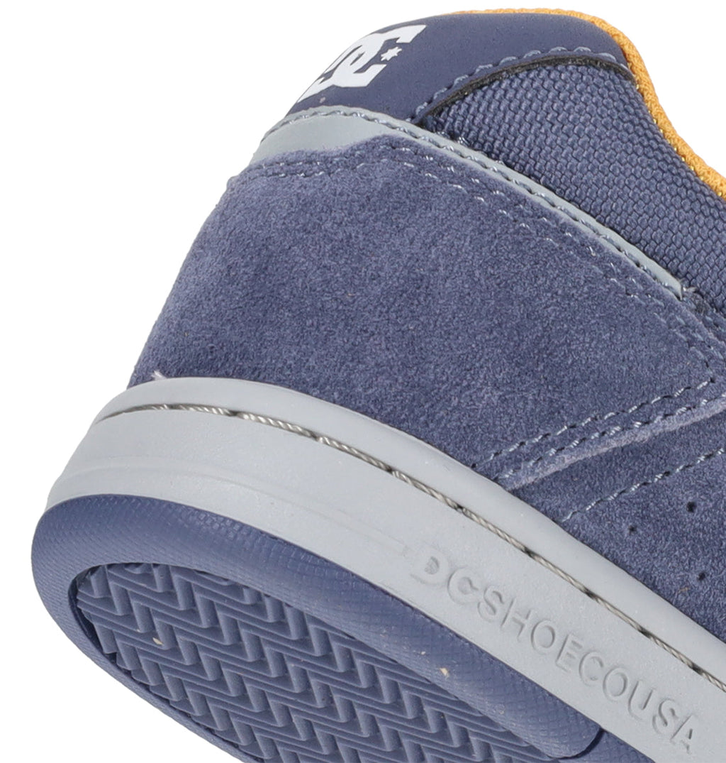 Close-up of the heel of a blue DC Shoes Kids’ Stag Shoe, featuring a gray sole with textured tread and visible suede and mesh materials. Designed for everyday wear, this vulcanized shoe showcases the partially visible DC logo on the upper heel.