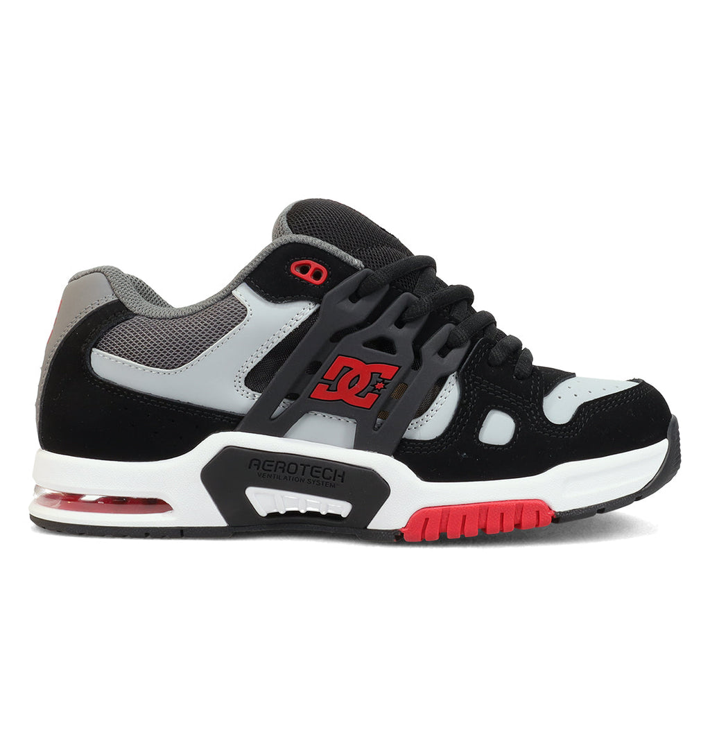 The Men’s AT-2 Shoes by DC Shoes feature a black, grey, and white design with bold red accents. They showcase the DC logo, a chunky lacing system, and a supportive TPU cage for enhanced stability. The midsole includes the AEROTECH ventilation system for breathability, while cushioned impact comfort and a red outsole provide superior grip.
