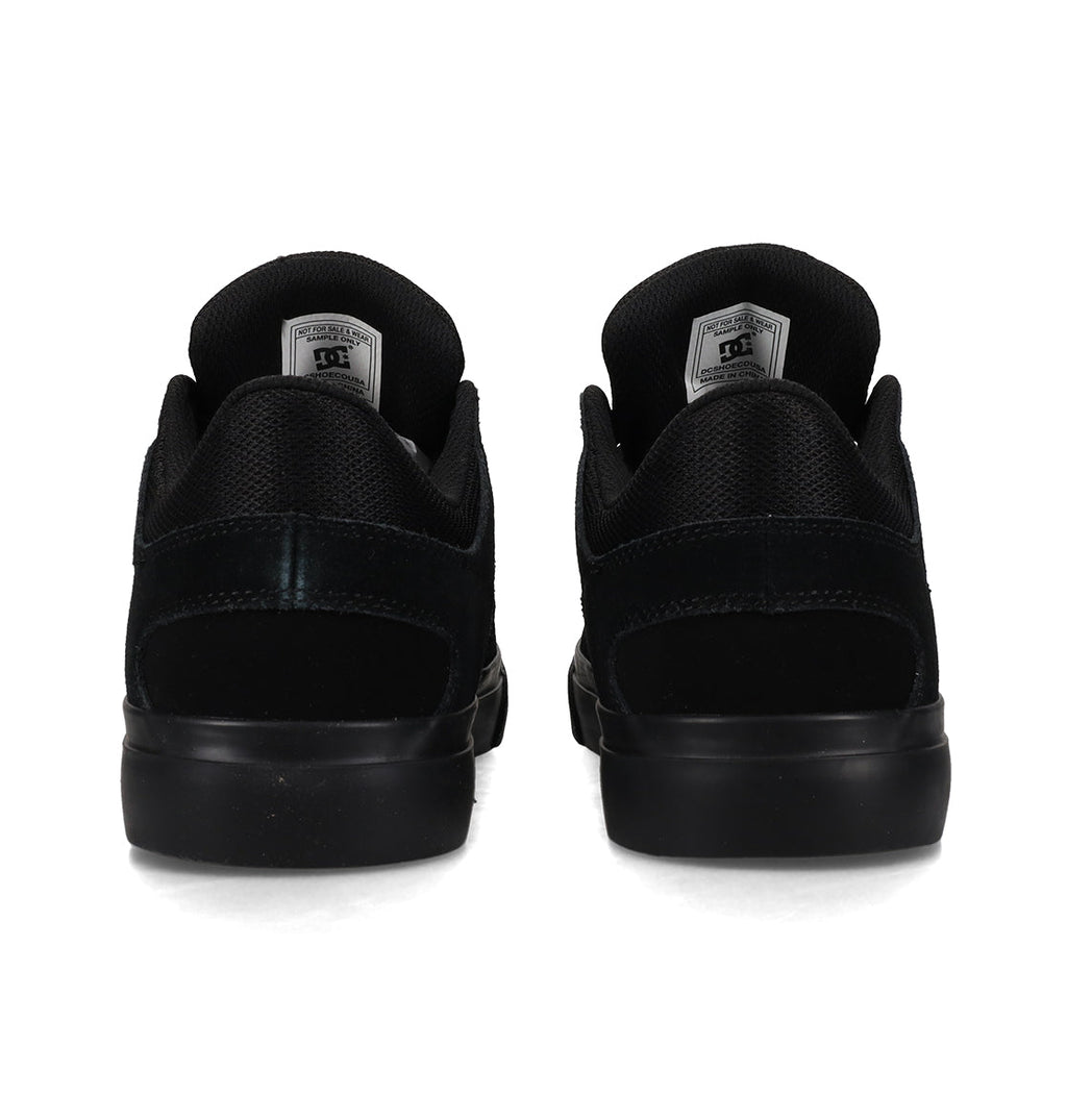A pair of DC Shoes Men’s Onyx Shoes in black, shown from the back, showcases a minimal silhouette with matching black soles and a DC logo tag on each tongue—an everyday vulc style crafted for both comfort and style.