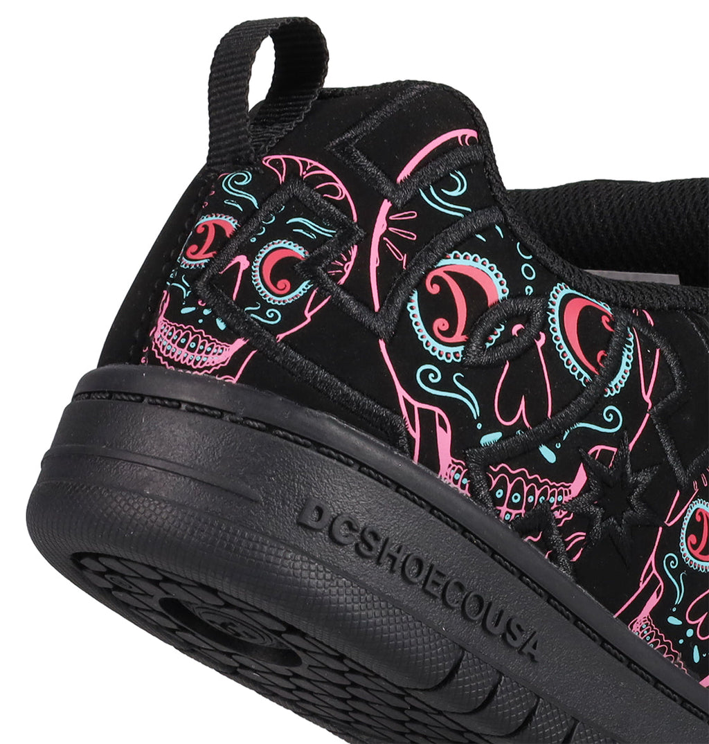 Close-up of the Kids’ Court Graffik Day Of The Dead sneaker by DC Shoes, part of the Day of the Dead Collection. This black shoe features colorful neon skull designs in pink, blue, and teal inspired by Día de los Muertos. The sole is printed with “DCSHOECOUSA,” and the design includes a heel pull tab and visible side stitching.