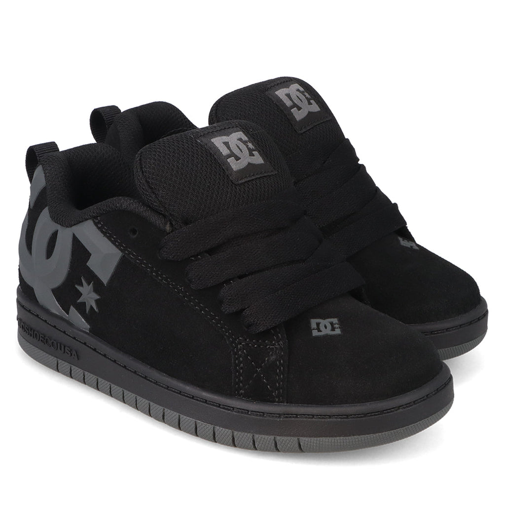 A pair of Kids’ Court Graffik Beveled Special Edition Shoes by DC Shoes in black, featuring thick black laces, a gray beveled TPR rubber logo on the side and tongue, and gray outsoles. The shoes showcase signature DC branding and are displayed side by side at an angle on a white background.