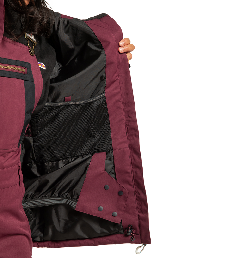 A person holds open the DC Shoes Women’s Liberate Insulated Jacket in maroon, revealing its black mesh inner pocket, cozy lining, and secure snap closures—ideal for winter adventures. Their face is hidden against a plain white background.
