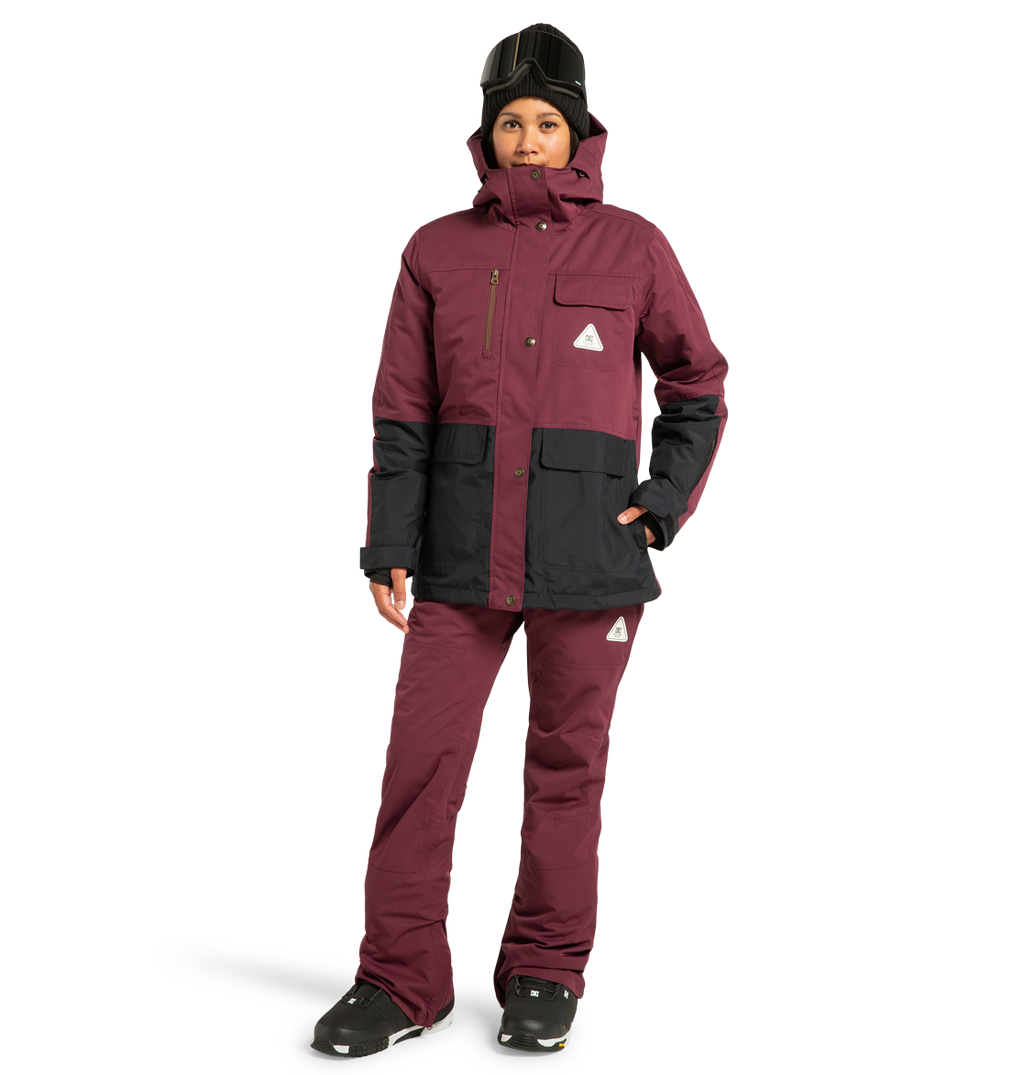 Wearing the DC Shoes Women’s Liberate Insulated Jacket in maroon and black, paired with matching maroon snow pants, black gloves, black boots, and a black helmet with goggles, this person stands against a plain white background—perfectly outfitted for any winter adventure with reliable weather protection.