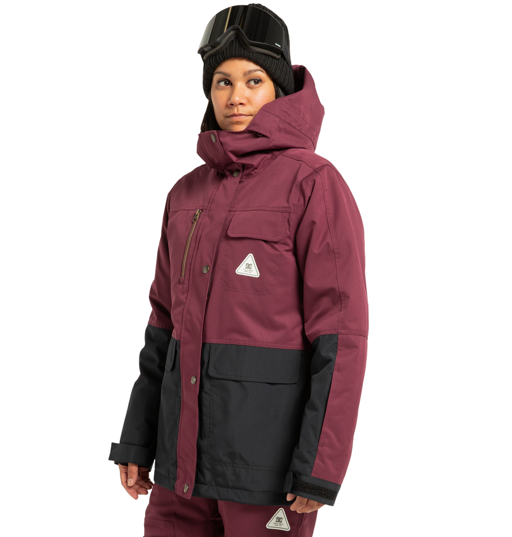 A person wearing a maroon and black DC Shoes Women’s Liberate Insulated Jacket, matching pants, a black beanie, and ski goggles stands against a plain white background—prepared for any winter adventure with reliable weather defense.