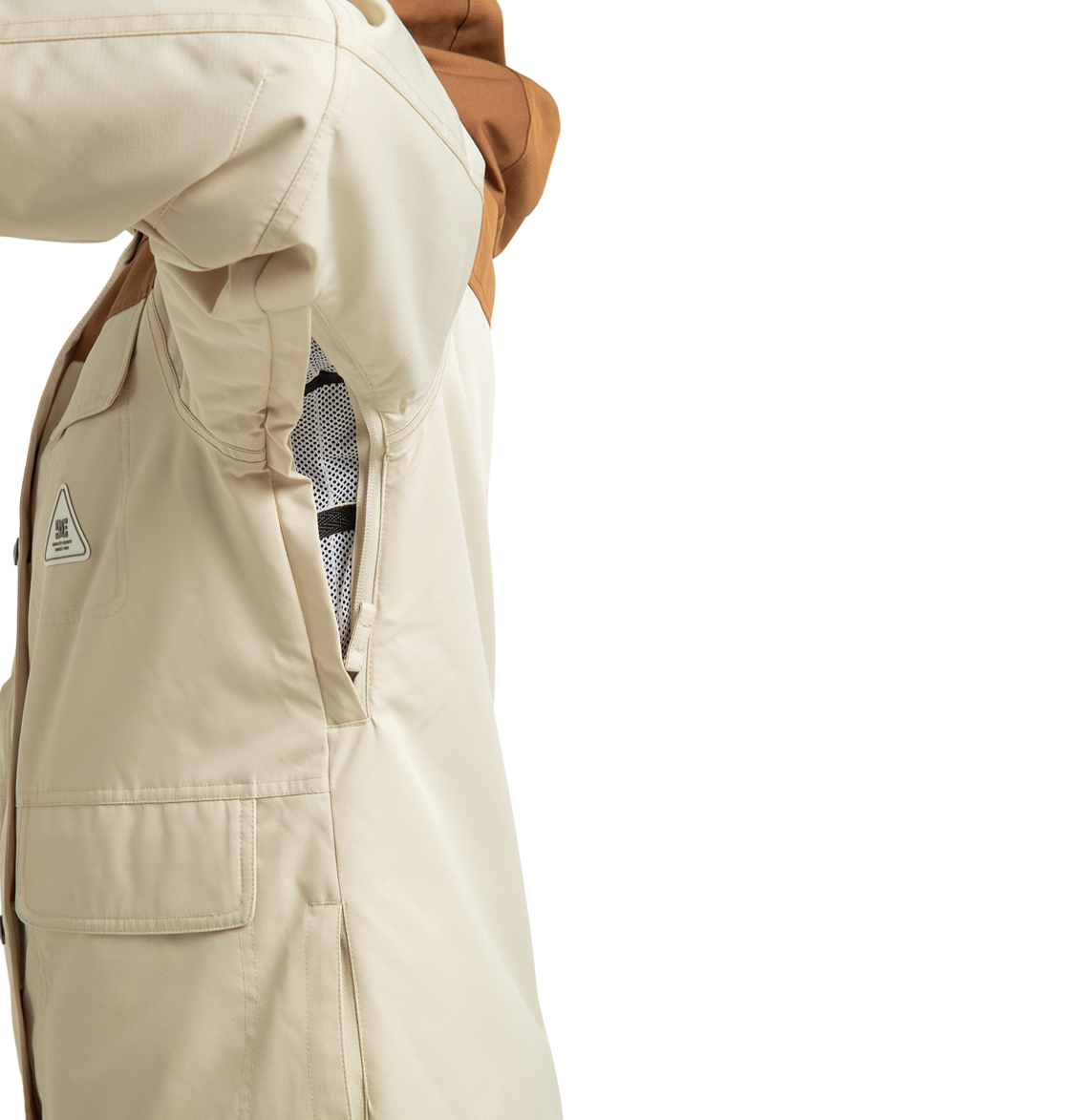 A close-up of a beige DC Shoes Women’s Liberate Insulated Jacket with a brown hood highlights an unzipped side vent, revealing the mesh lining inside. Crafted from recycled polyester, this jacket is ideal for winter adventures. The wearer has their arm raised, showing a flap pocket on the side.