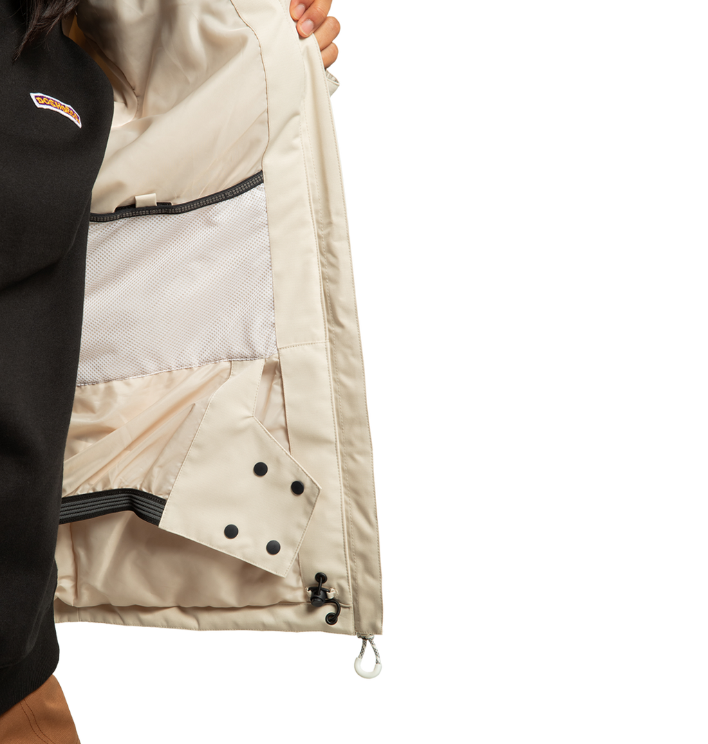 A person holds open a light beige DC Shoes Women’s Liberate Insulated Jacket, revealing interior mesh and zippered pockets, black snap buttons, and drawstring details—perfect for any winter adventure. Part of their black shirt and brown pants are visible. This jacket is thoughtfully crafted from recycled polyester.