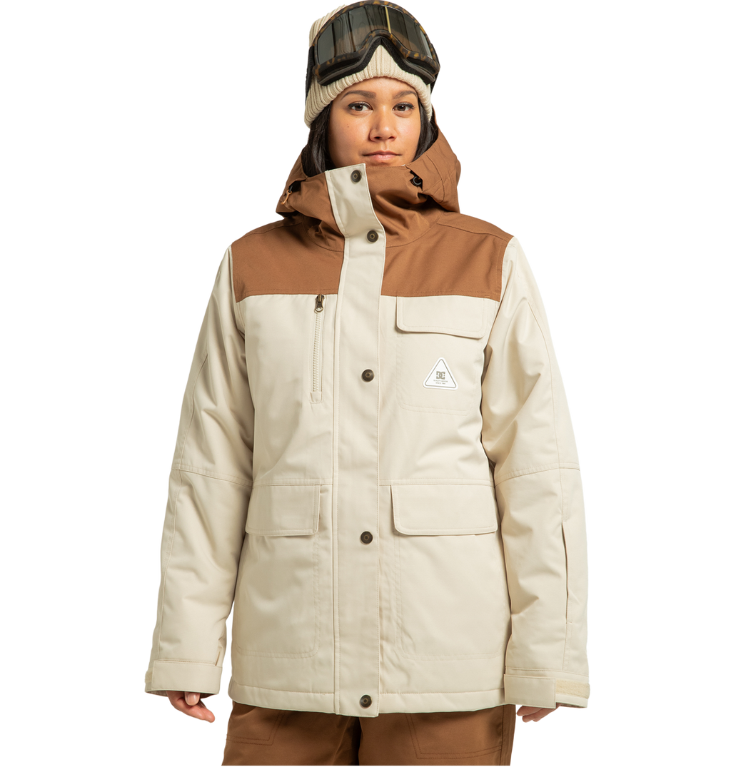 A person stands against a plain white background ready for any winter adventure, dressed in the DC Shoes Women’s Liberate Insulated Jacket—crafted from recycled polyester in beige and brown—paired with brown pants, a beanie, and ski goggles on their head.