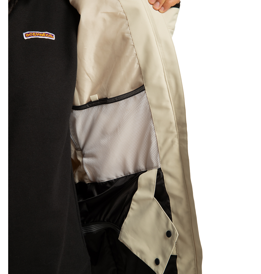 A person in a black DC Shoes Women’s Cruiser Insulated Jacket, made from recycled polyester and featuring weather defense technology, holds open a beige coat to reveal its light gray interior lining and mesh pocket. The DOCKHOUSE logo is visible on the black jacket.
