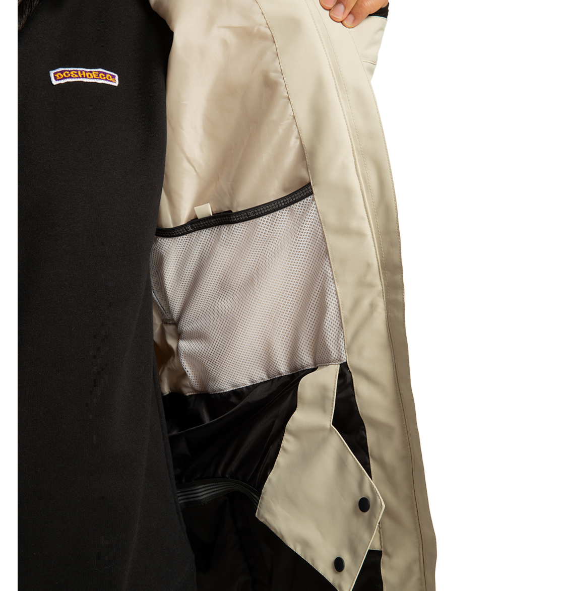 A person in a black DC Shoes Women’s Cruiser Insulated Jacket, made from recycled polyester and featuring weather defense technology, holds open a beige coat to reveal its light gray interior lining and mesh pocket. The DOCKHOUSE logo is visible on the black jacket.