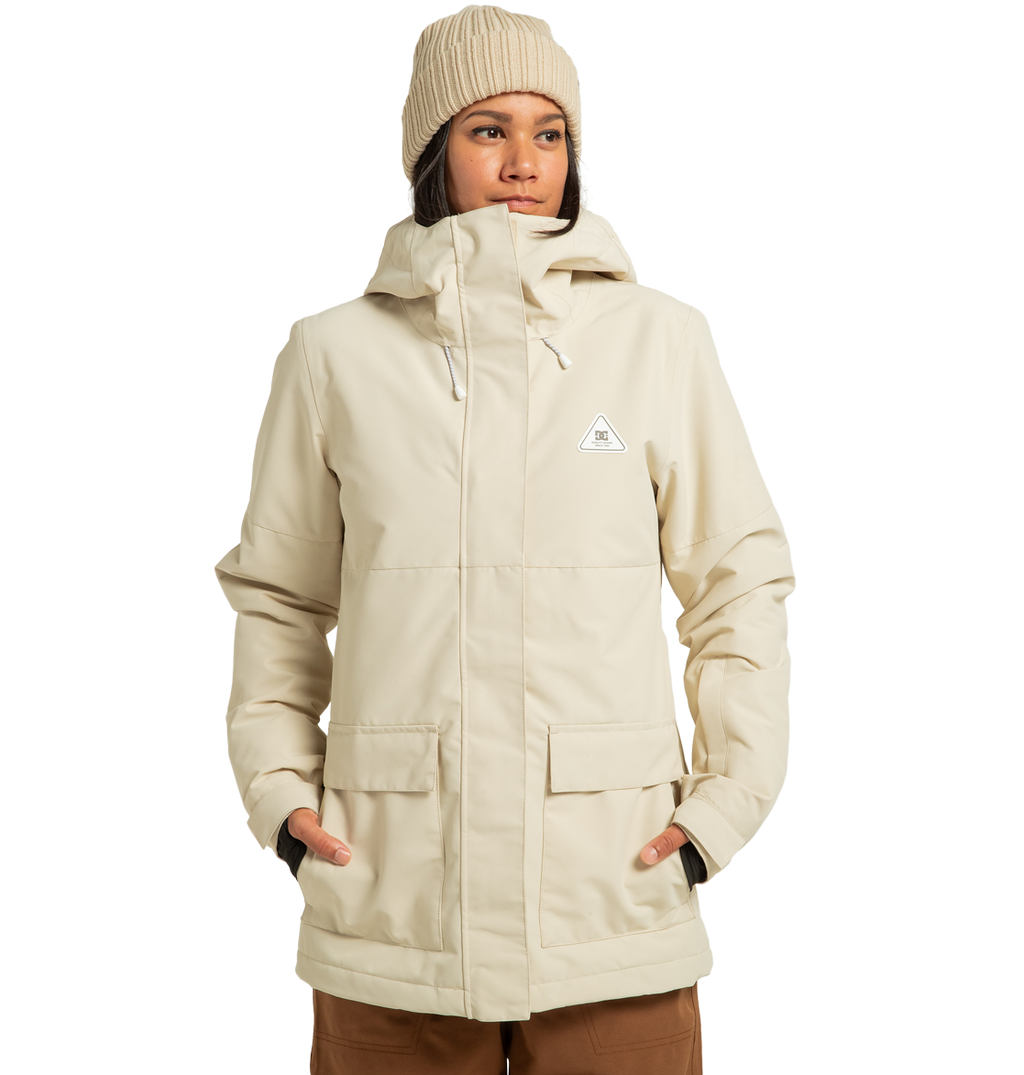 A person stands against a plain white background with hands in the pockets of the DC Shoes Women’s Cruiser Insulated Jacket—a beige, hooded winter jacket paired with a matching knit beanie. Made from recycled polyester and enhanced with weather defense technology.