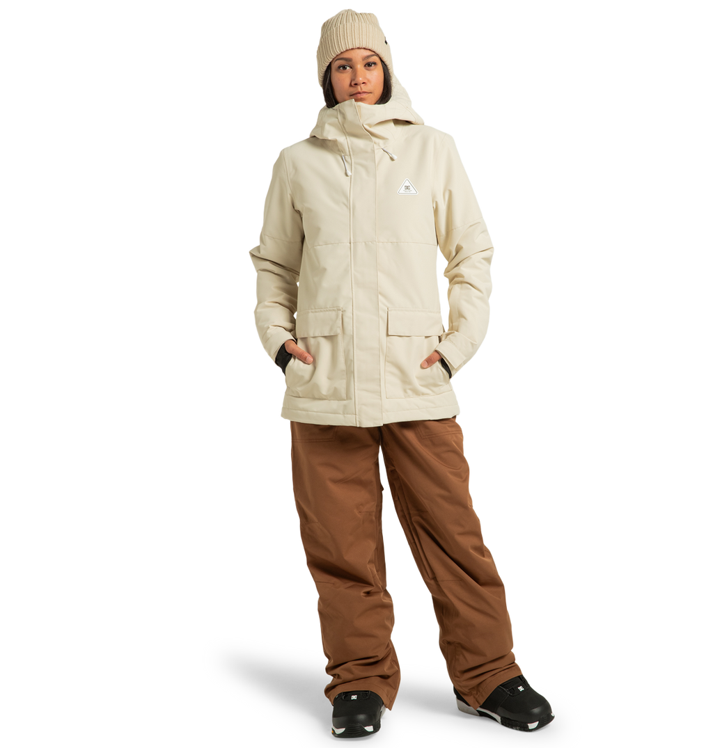 A woman stands with her hands in the pockets of her DC Shoes Women’s Cruiser Insulated Jacket, crafted from recycled polyester and featuring advanced weather defense technology, paired with brown pants, black boots, and a beige beanie against a plain white background.