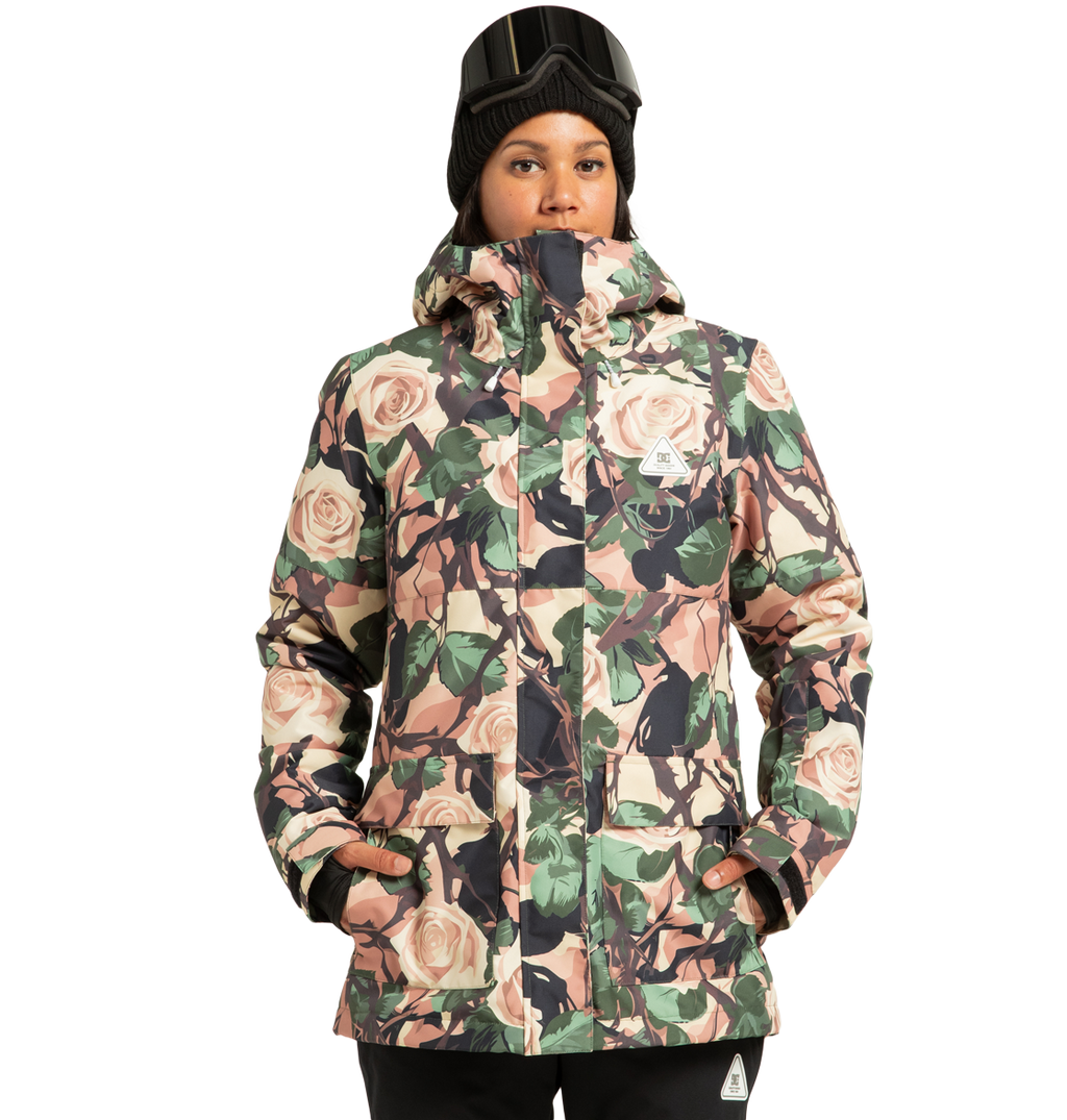 A person stands with hands in pockets against a plain background, wearing the DC Shoes Women’s Cruiser Insulated Snow Jacket with a floral pattern, paired with a black beanie and ski goggles resting on their forehead.