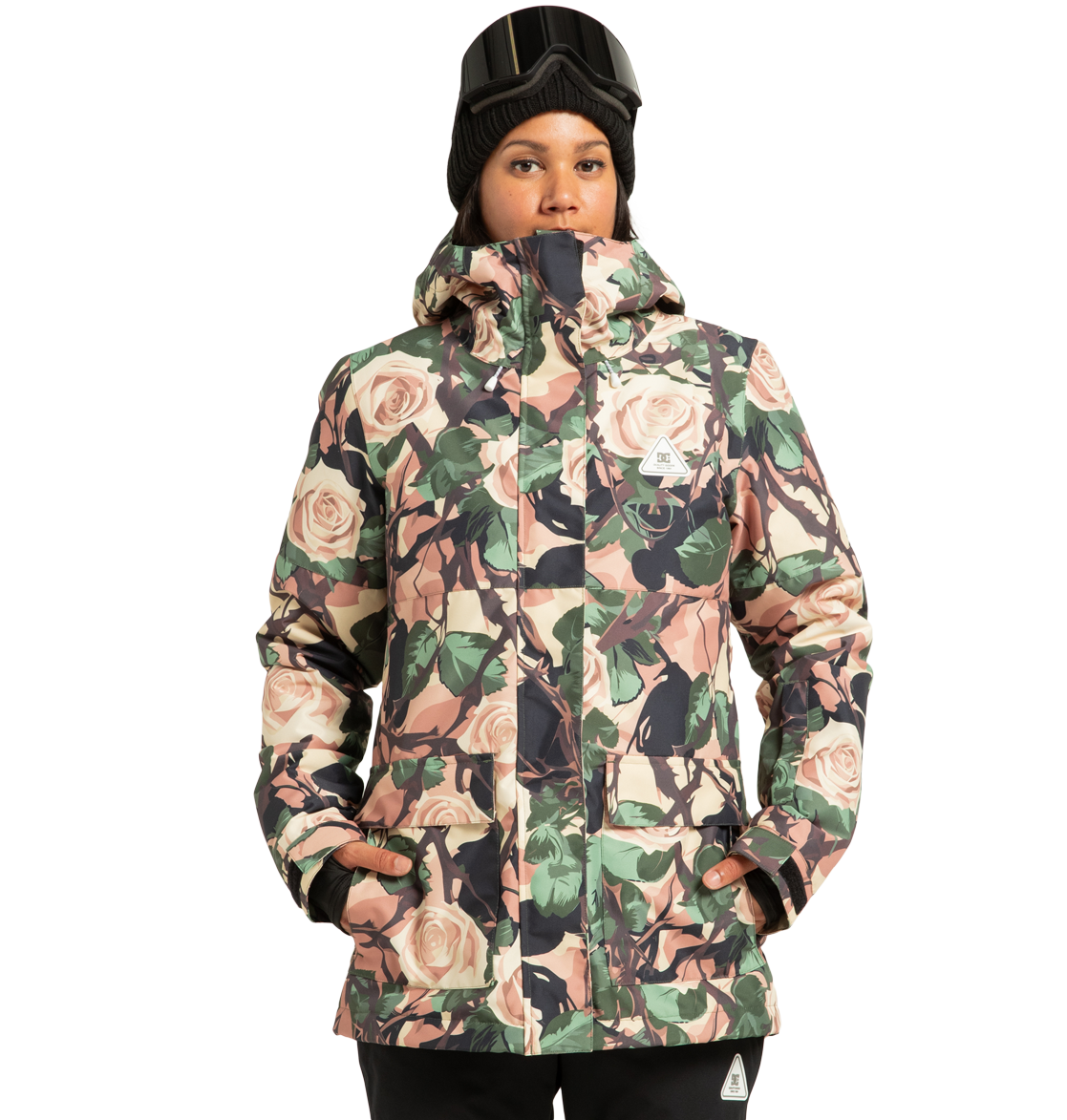 A person stands with hands in pockets against a plain background, wearing the DC Shoes Women’s Cruiser Insulated Snow Jacket with a floral pattern, paired with a black beanie and ski goggles resting on their forehead.