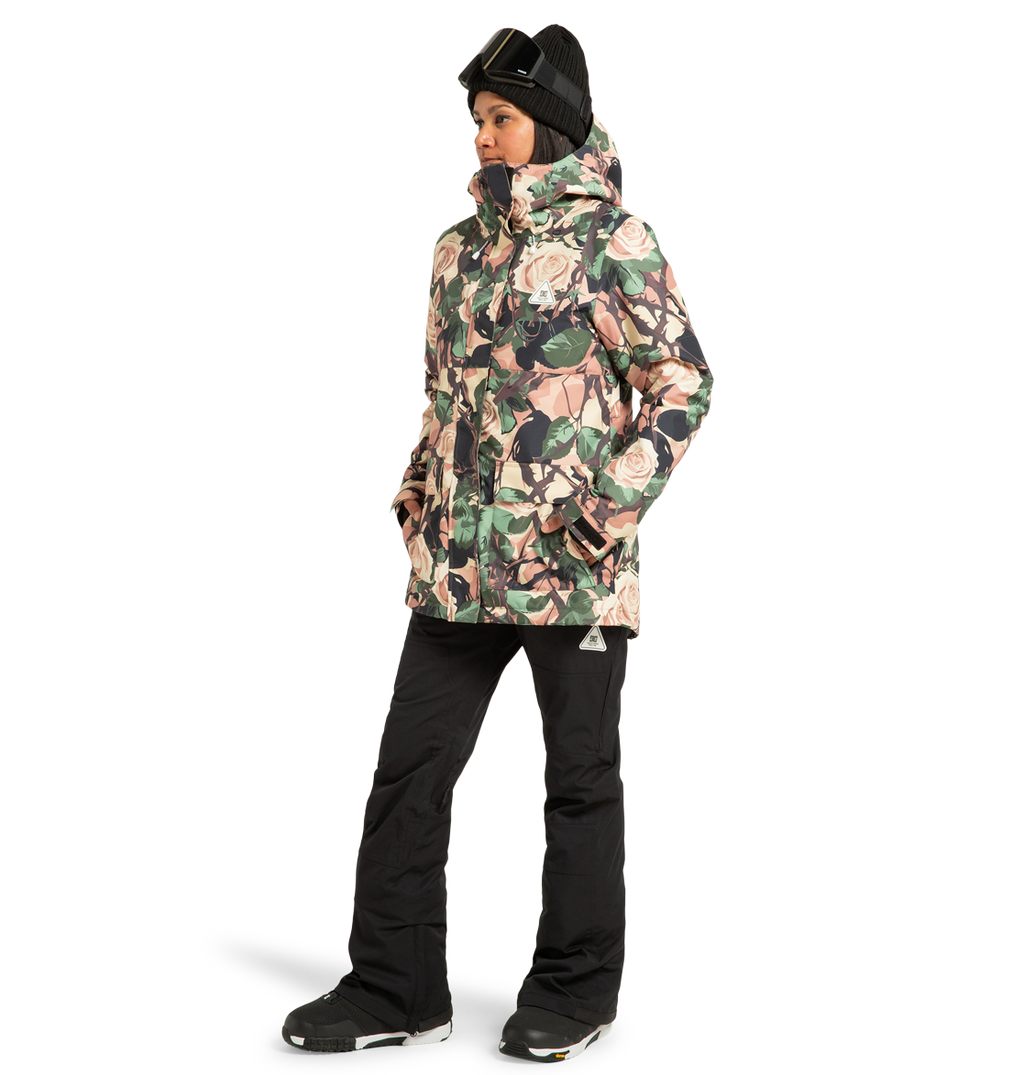 A person wearing the DC Shoes Women’s Cruiser Insulated Snow Jacket, featuring a floral pattern made from recycled polyester, paired with black snow pants, black gloves, a black beanie, and ski goggles, stands with hands in pockets against a plain white background.