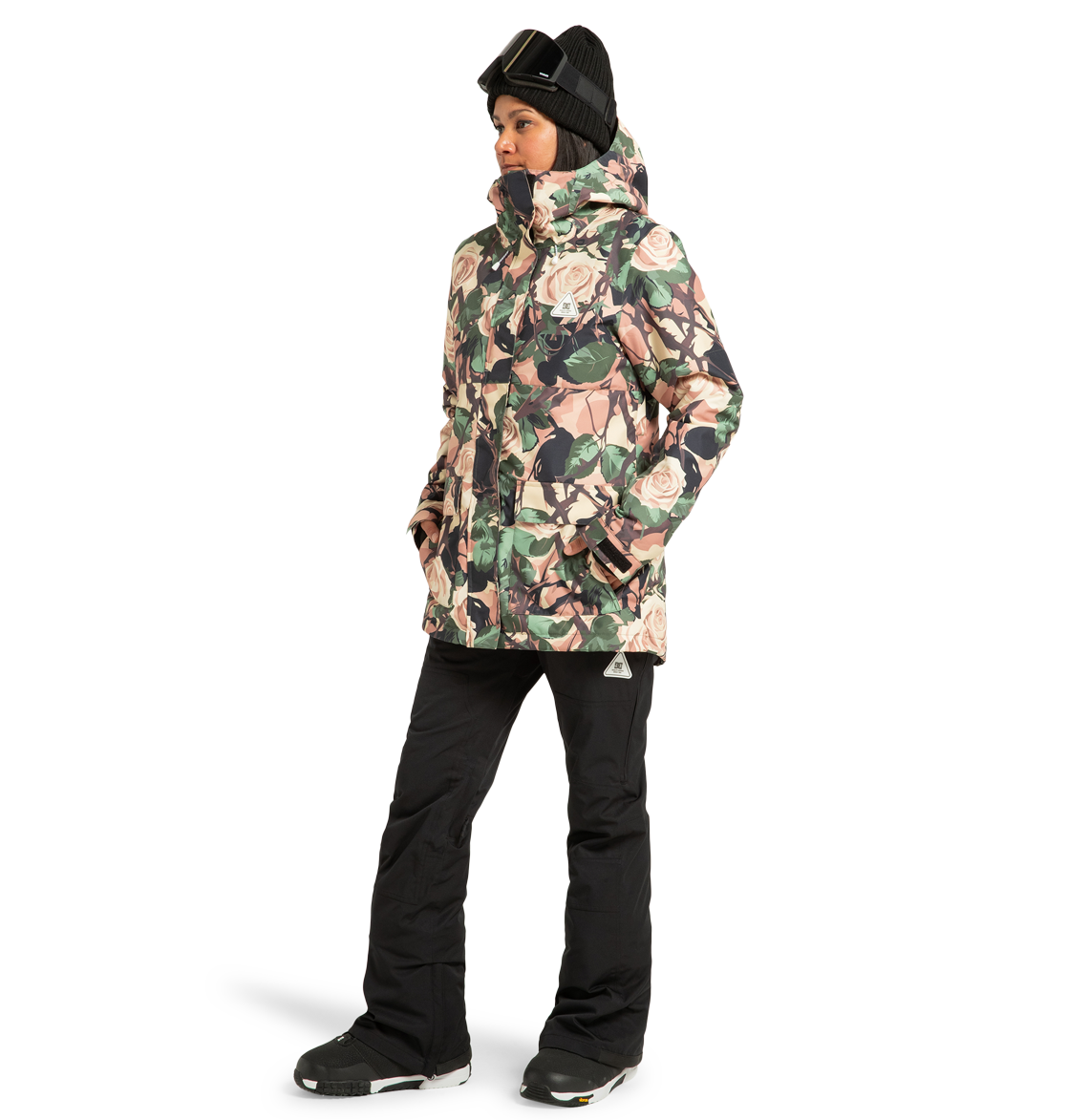 A person wearing the DC Shoes Women’s Cruiser Insulated Snow Jacket, featuring a floral pattern made from recycled polyester, paired with black snow pants, black gloves, a black beanie, and ski goggles, stands with hands in pockets against a plain white background.