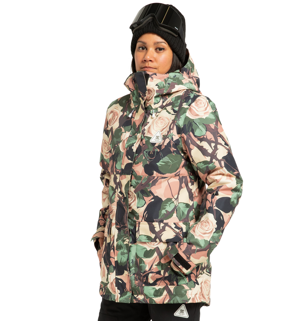A person stands with hands in pockets, looking at the camera against a plain white background, dressed in a beanie, ski goggles, and the DC Shoes Women’s Cruiser Insulated Snow Jacket featuring a floral pattern and made from recycled polyester.