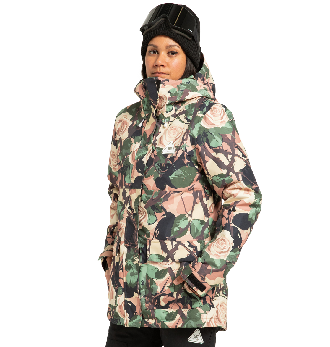 A person stands with hands in pockets, looking at the camera against a plain white background, dressed in a beanie, ski goggles, and the DC Shoes Women’s Cruiser Insulated Snow Jacket featuring a floral pattern and made from recycled polyester.