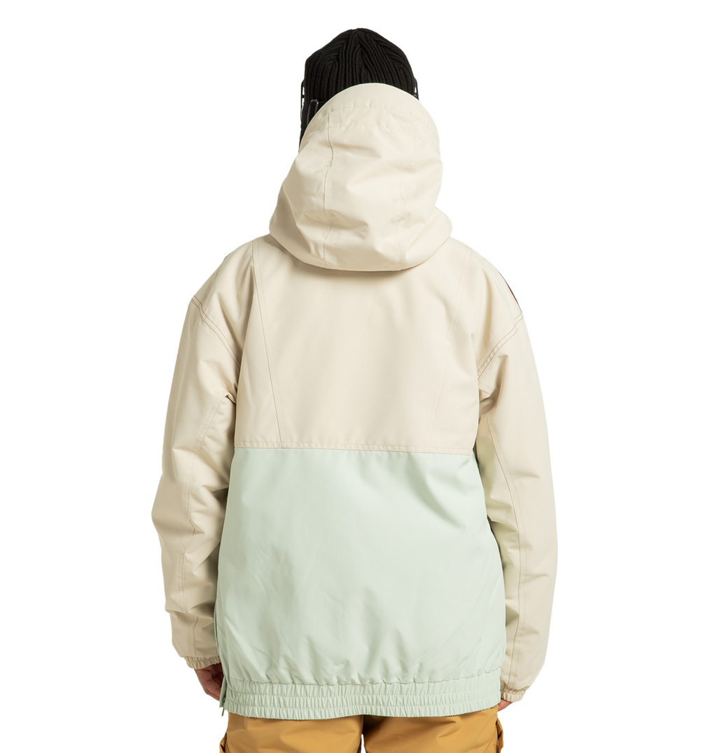 A person stands with their back to the camera against a plain white background, wearing the DC Shoes Women’s Chalet Anorak Insulated Jacket in a cream and light green color-blocked design, paired with brown pants and a black beanie.