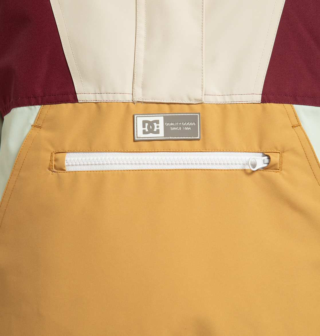 Close-up of the DC Shoes Women’s Chalet Anorak Insulated Jacket, showcasing mustard yellow fabric, a white zippered pocket, and a small metal label near the top that reads “DC QUALITY GOODS SINCE 1994,” perfect for outdoor adventures.