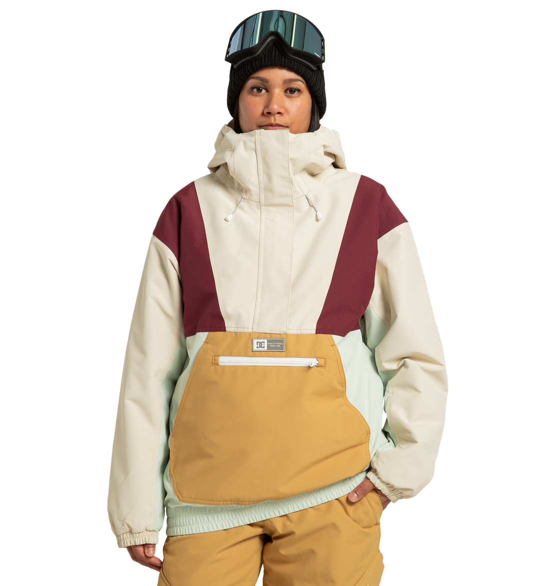 A person stands against a plain white background, ready for any outdoor adventure, wearing the DC Shoes Women’s Chalet Anorak Insulated Jacket—crafted from recycled polyester with beige, maroon, mint, and mustard colorblocking—paired with tan pants, a black beanie, and ski goggles.