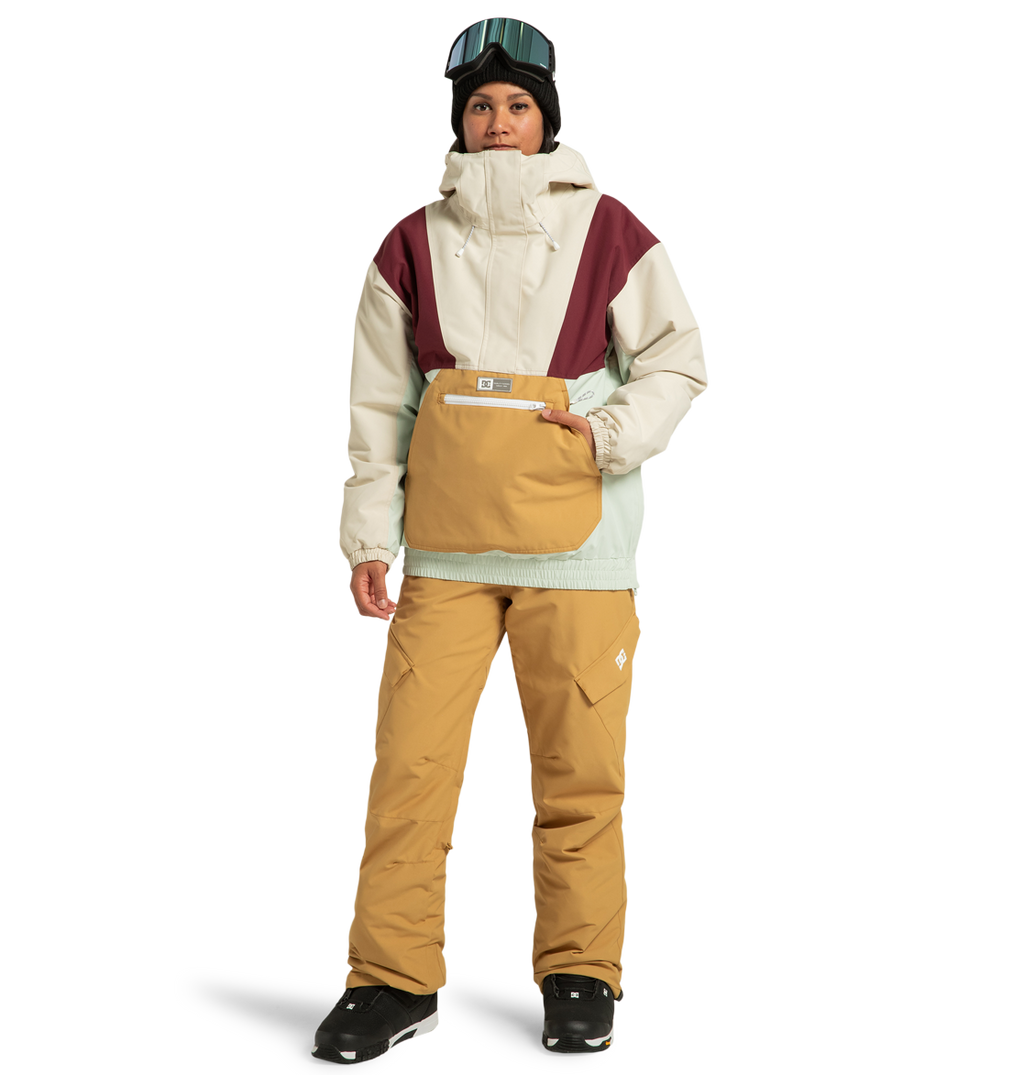 A person is wearing the DC Shoes Women’s Chalet Anorak Insulated Jacket in beige, burgundy, and mint—crafted from recycled polyester—paired with tan pants, black boots, and a black beanie topped with goggles, standing against a white background.