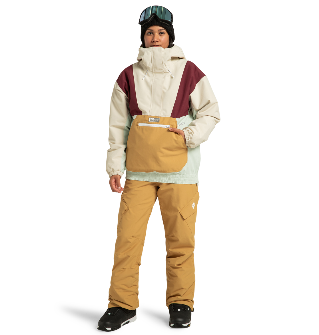 A person is wearing the DC Shoes Women’s Chalet Anorak Insulated Jacket in beige, burgundy, and mint—crafted from recycled polyester—paired with tan pants, black boots, and a black beanie topped with goggles, standing against a white background.