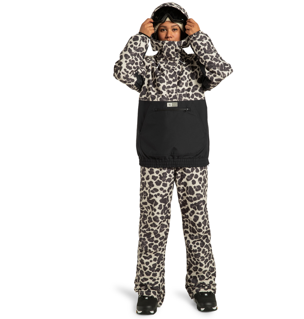 A woman wearing the DC Shoes Women’s Chalet Anorak Insulated Jacket in a black and white leopard print, paired with black boots, stands against a plain white background. Crafted from recycled polyester for warmth and sustainability, the stylish jacket is complemented by her ski goggles, which she adjusts atop her head.