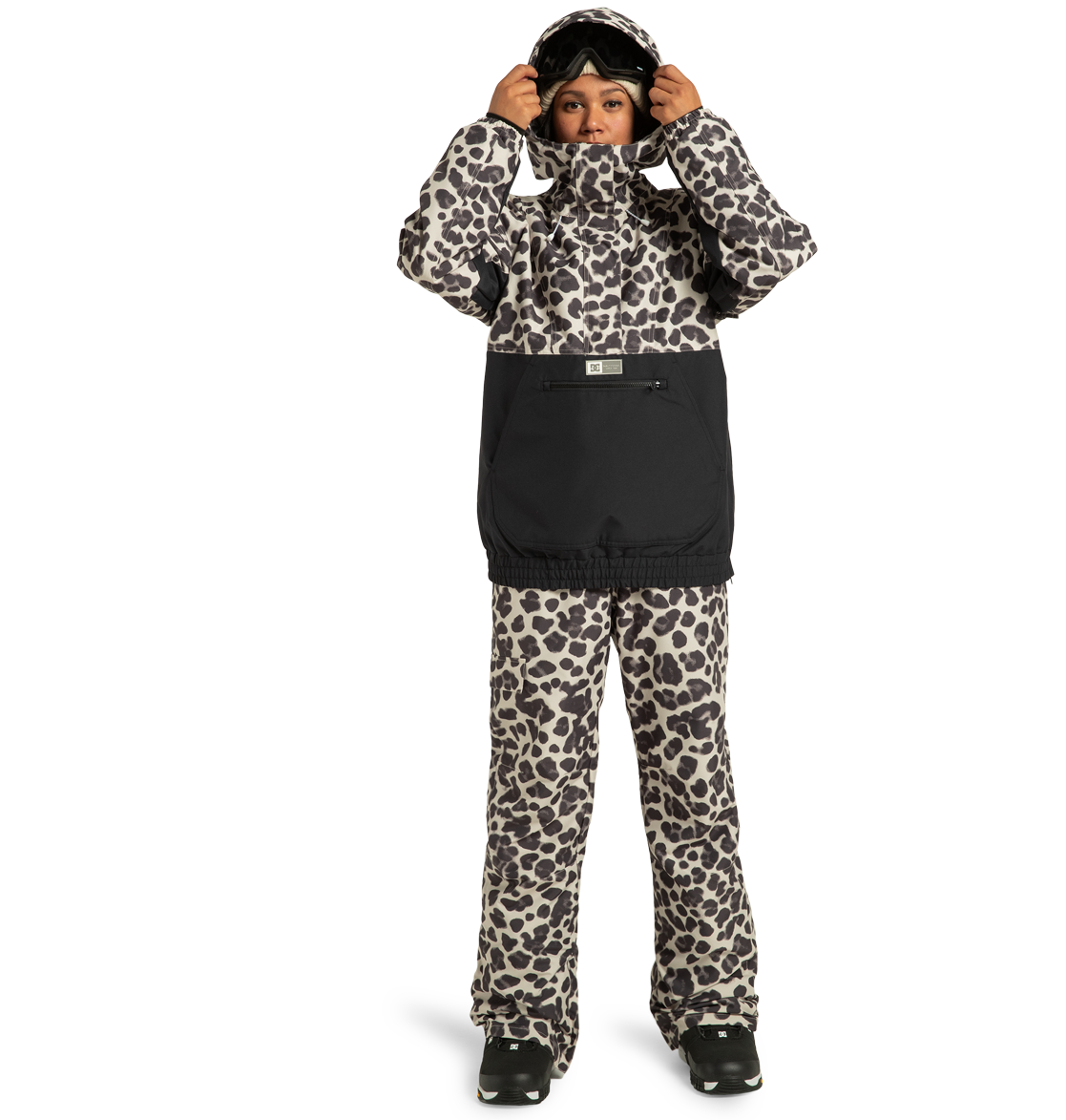 A woman wearing the DC Shoes Women’s Chalet Anorak Insulated Jacket in a black and white leopard print, paired with black boots, stands against a plain white background. Crafted from recycled polyester for warmth and sustainability, the stylish jacket is complemented by her ski goggles, which she adjusts atop her head.
