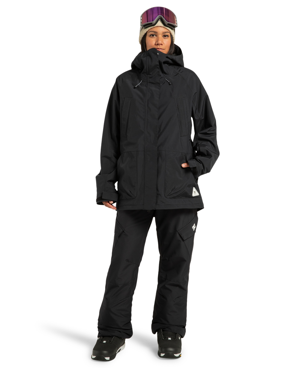 A person wearing DC Shoes Women’s Nonchalant Insulated Snow Pants in recycled polyester, a black winter jacket, black boots, a gray knit hat, and ski goggles pushed up on their forehead stands against a plain white background.