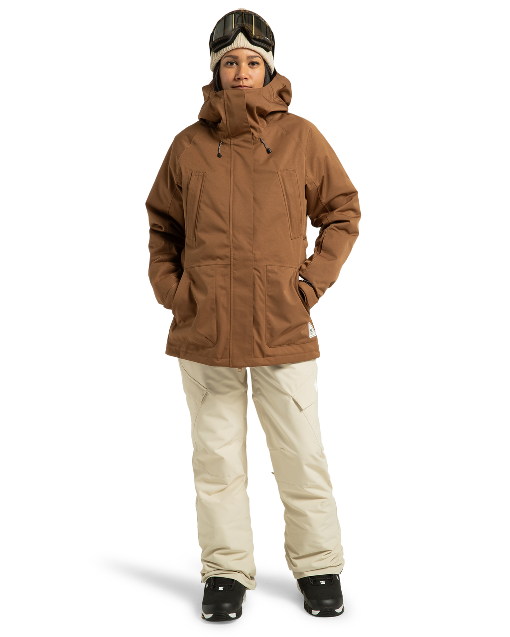 A person wearing a brown hooded winter jacket, DC Shoes Women’s Nonchalant Insulated Snow Pants in beige made from recycled polyester, black gloves, black boots, and ski goggles stands facing forward with hands in jacket pockets against a plain white background.