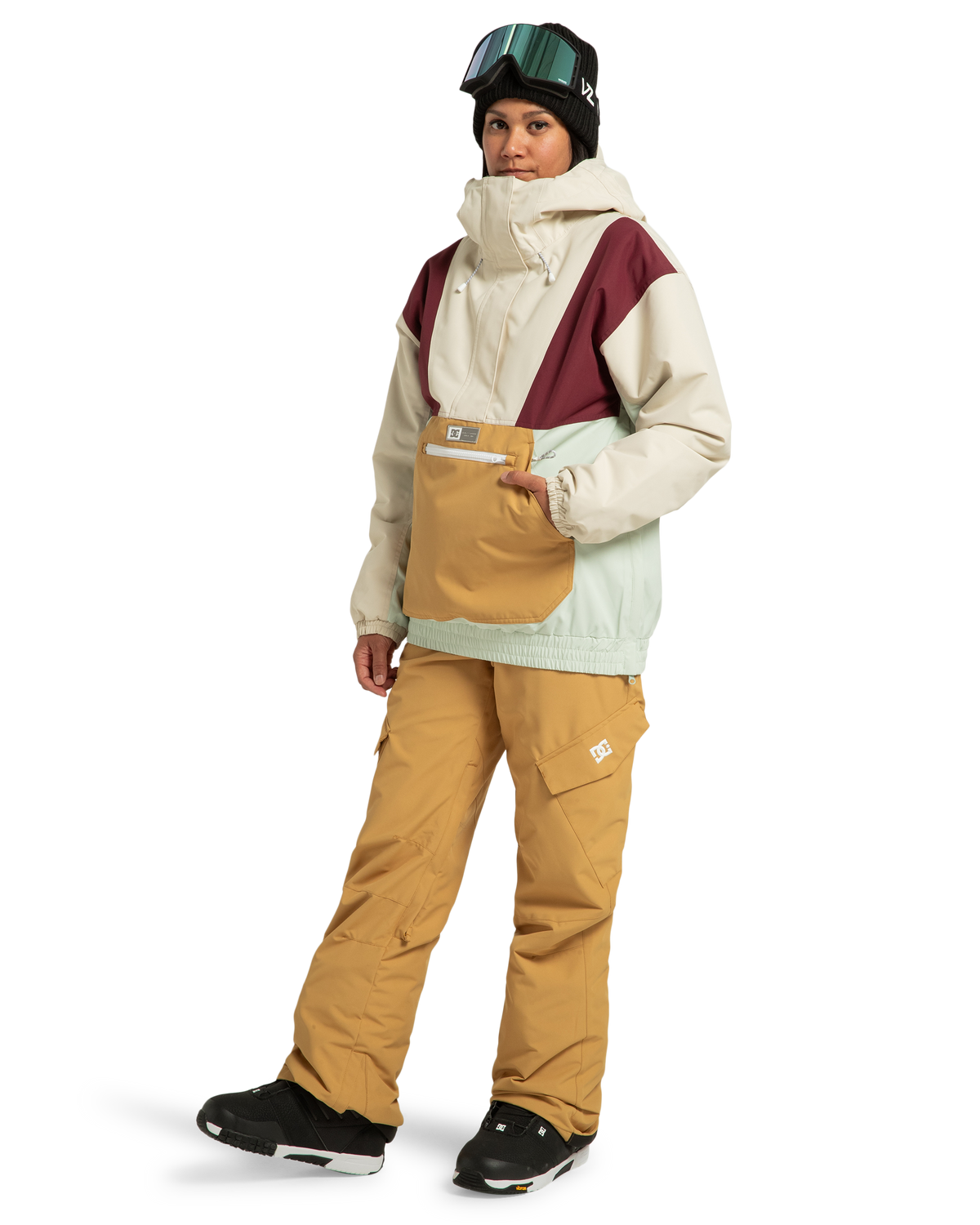 A person in snow gear wears a cream, mint, and burgundy jacket paired with DC Shoes Women’s Nonchalant Insulated Snow Pants in mustard yellow—crafted from recycled polyester—along with black boots, black gloves, a black beanie, and ski goggles, standing and facing forward against a white background.