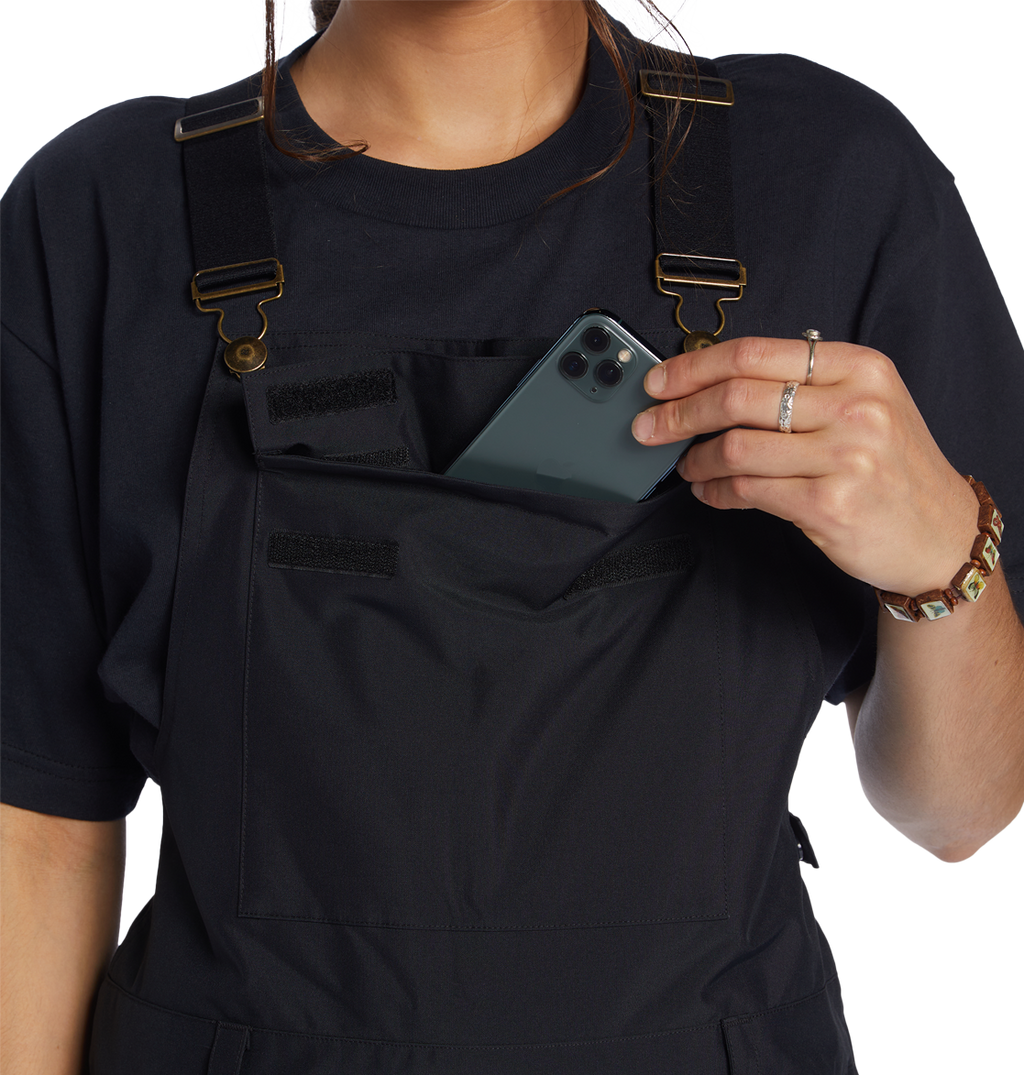 A person in a black shirt and DC Shoes Women’s Valiant Bib, crafted from recycled polyester, slips a smartphone into the front chest pocket of the overalls. The image focuses on the upper body, emphasizing the bib’s Weather Defense 10 protection.