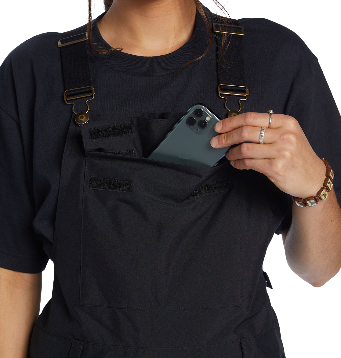 A person in a black shirt and DC Shoes Women’s Valiant Bib, crafted from recycled polyester, slips a smartphone into the front chest pocket of the overalls. The image focuses on the upper body, emphasizing the bib’s Weather Defense 10 protection.