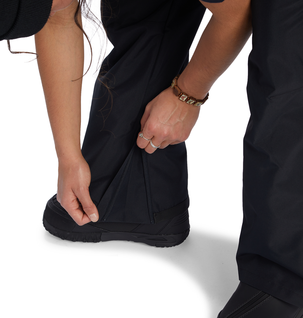 A person zips up the lower leg of the DC Shoes Women’s Valiant Bib, made from recycled polyester, over a black boot. They are wearing a brown bracelet and a ring on their left hand.