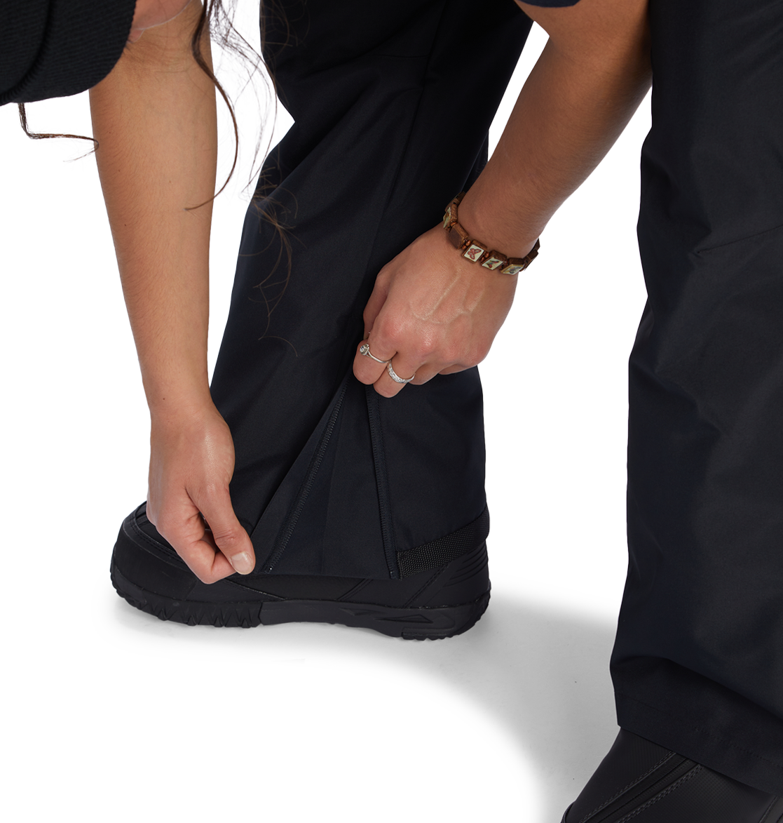A person zips up the lower leg of the DC Shoes Women’s Valiant Bib, made from recycled polyester, over a black boot. They are wearing a brown bracelet and a ring on their left hand.