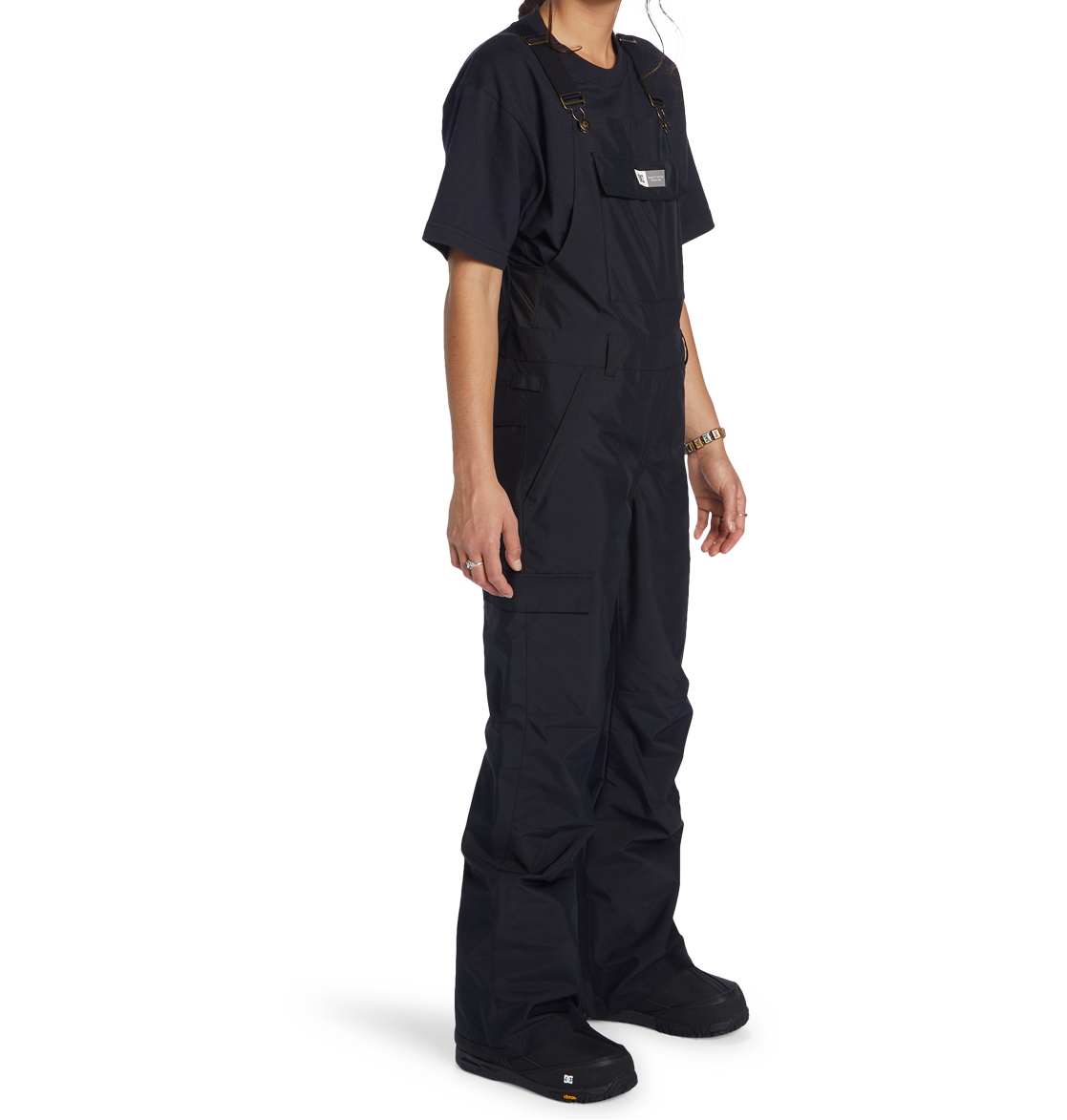 A person stands facing slightly right against a plain white background, wearing a black T-shirt and the DC Shoes Women’s Valiant Bib with pockets, crafted from recycled polyester. Their face is not visible.