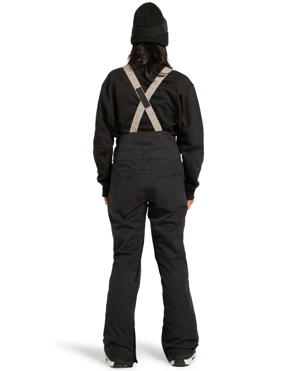 A person stands with their back to the camera against a plain white background, wearing the DC Shoes Women’s Crusade Shell Bib snow pants with suspenders, paired with a black beanie and black jacket—highlighting women’s adventure gear engineered for waterproof protection.