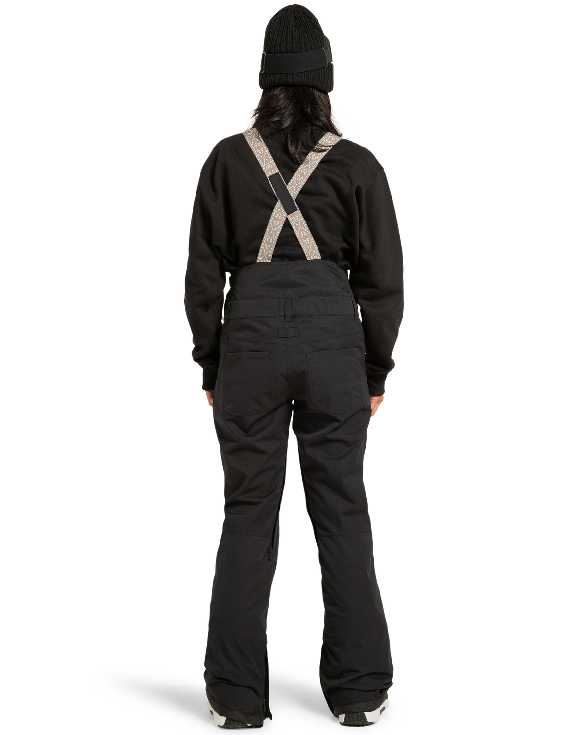 A person stands with their back to the camera against a plain white background, wearing the DC Shoes Women’s Crusade Shell Bib snow pants with suspenders, paired with a black beanie and black jacket—highlighting women’s adventure gear engineered for waterproof protection.