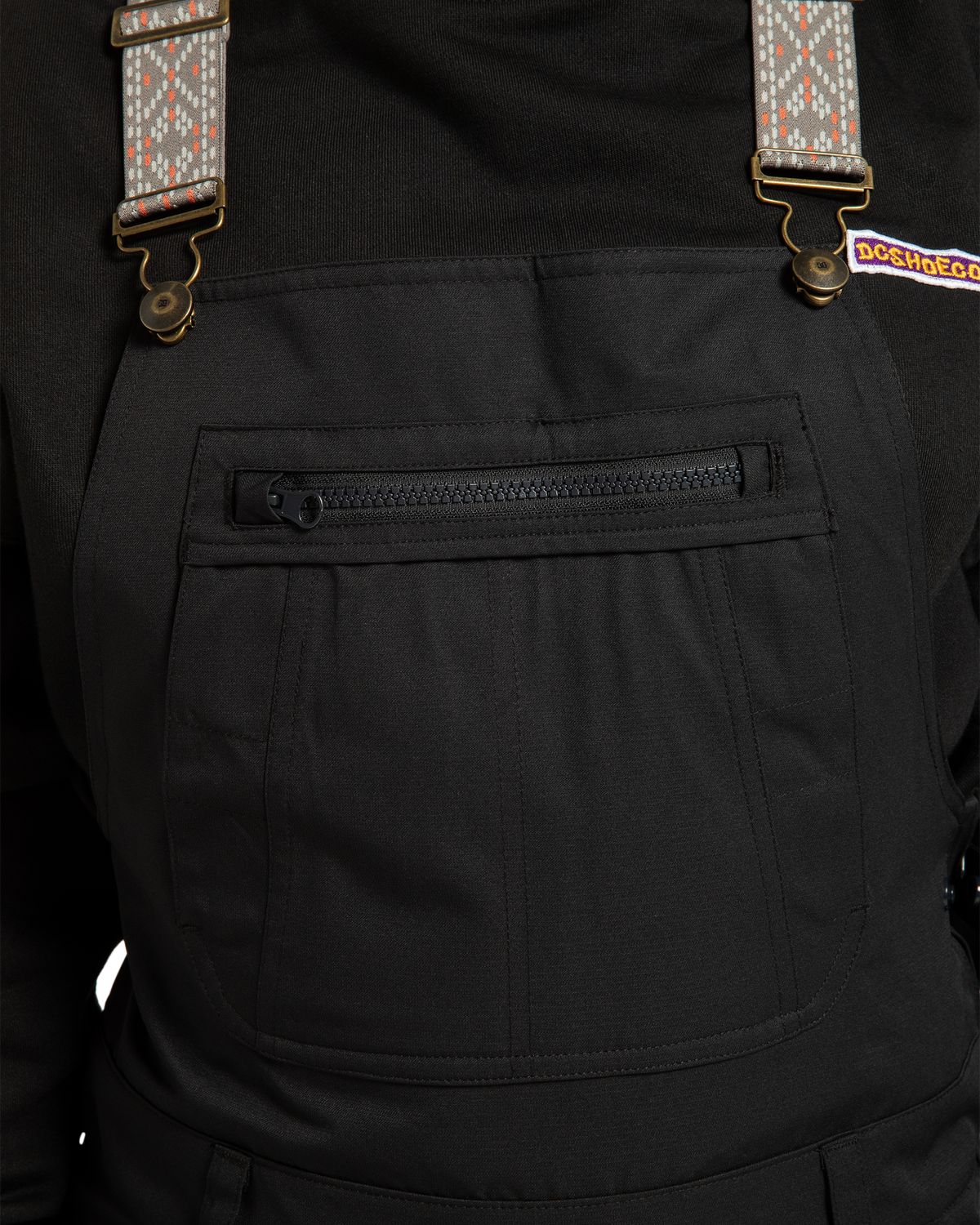 Close-up of a person wearing black DC Shoes Women’s Crusade Shell Bib overalls with patterned suspenders. These adventure-ready overalls feature a zippered front pocket and a small colorful DC Shoes logo on the upper right side, providing stylish waterproof protection for any journey.