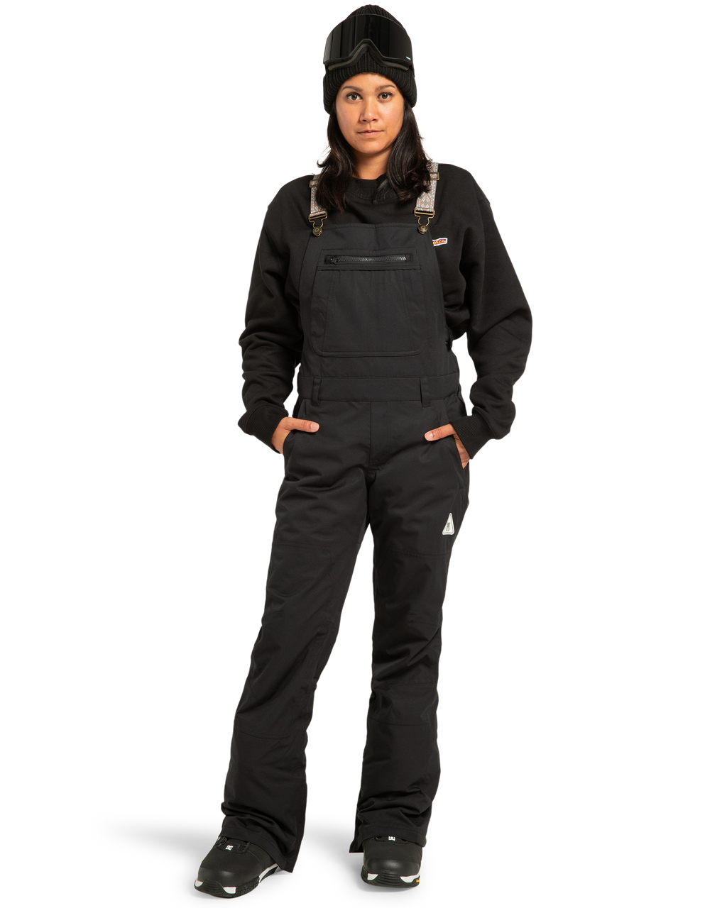 A person wearing the DC Shoes Women’s Crusade Shell Bib in black, paired with a black sweatshirt, black boots, beanie, and ski goggles—standing with hands in pockets against a white background—showcases women’s adventure gear engineered for ultimate waterproof protection.