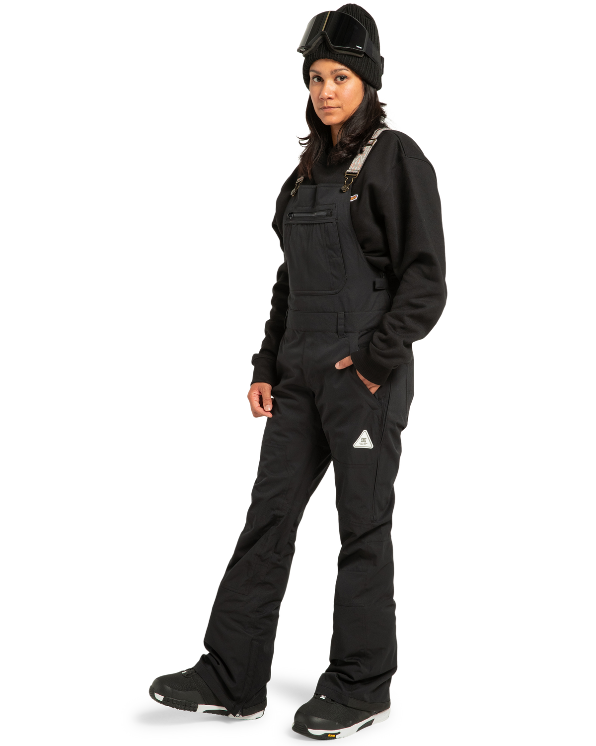 A person stands facing forward against a white background, wearing DC Shoes Women’s Crusade Shell Bib black snow overalls, paired with a black sweatshirt, black boots, a black beanie, and ski goggles; their arms are relaxed by their sides as they showcase waterproof women’s adventure gear.