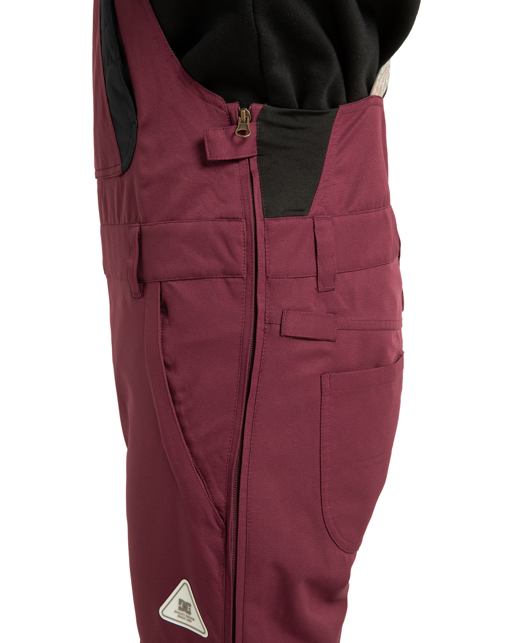 Side view close-up of a person wearing the maroon Women’s Crusade Shell Bib from DC Shoes—an eco-friendly ski bib featuring a zipper, belt loops, multiple pockets, and a small triangular logo on the thigh. This women’s snow bib is paired with a black top, offering both functional performance and stylish appeal.